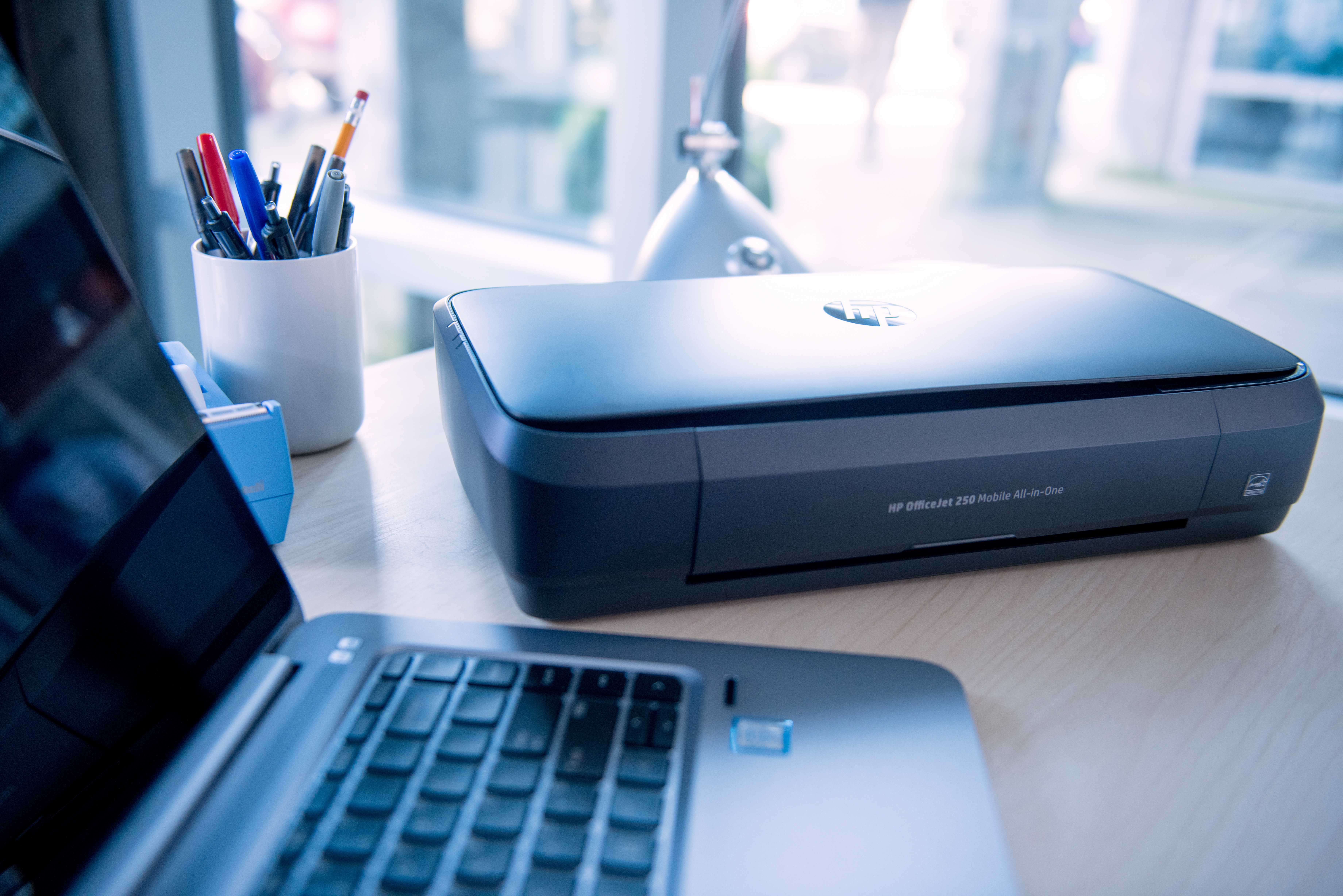 Ein Laptop, Drucker und Stiftehalter auf einem Holztisch. Draußen ist es hell.