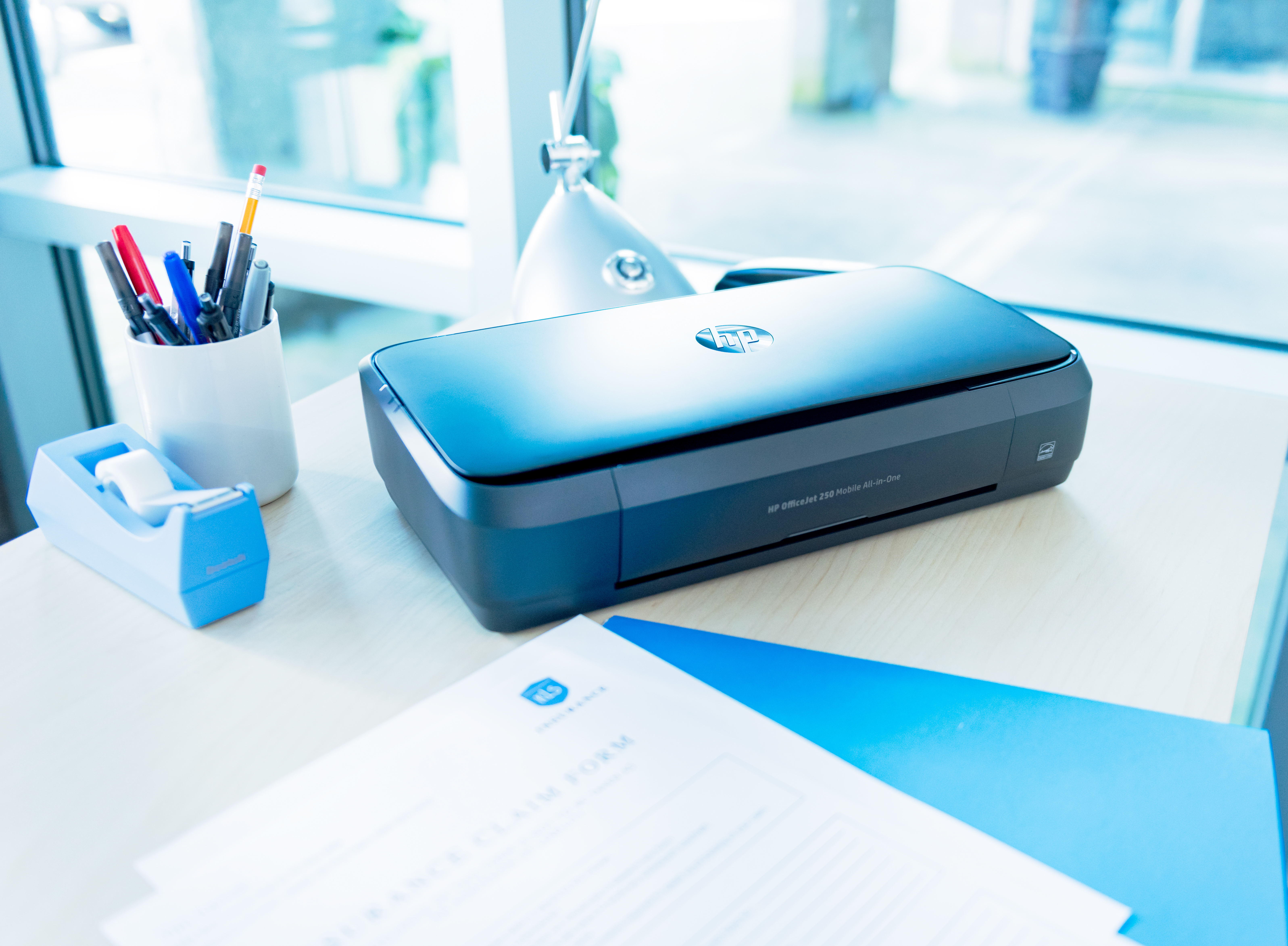 Ein schwarzer HP Drucker steht auf einem Schreibtisch neben einem Klebebandspender und Stiften.