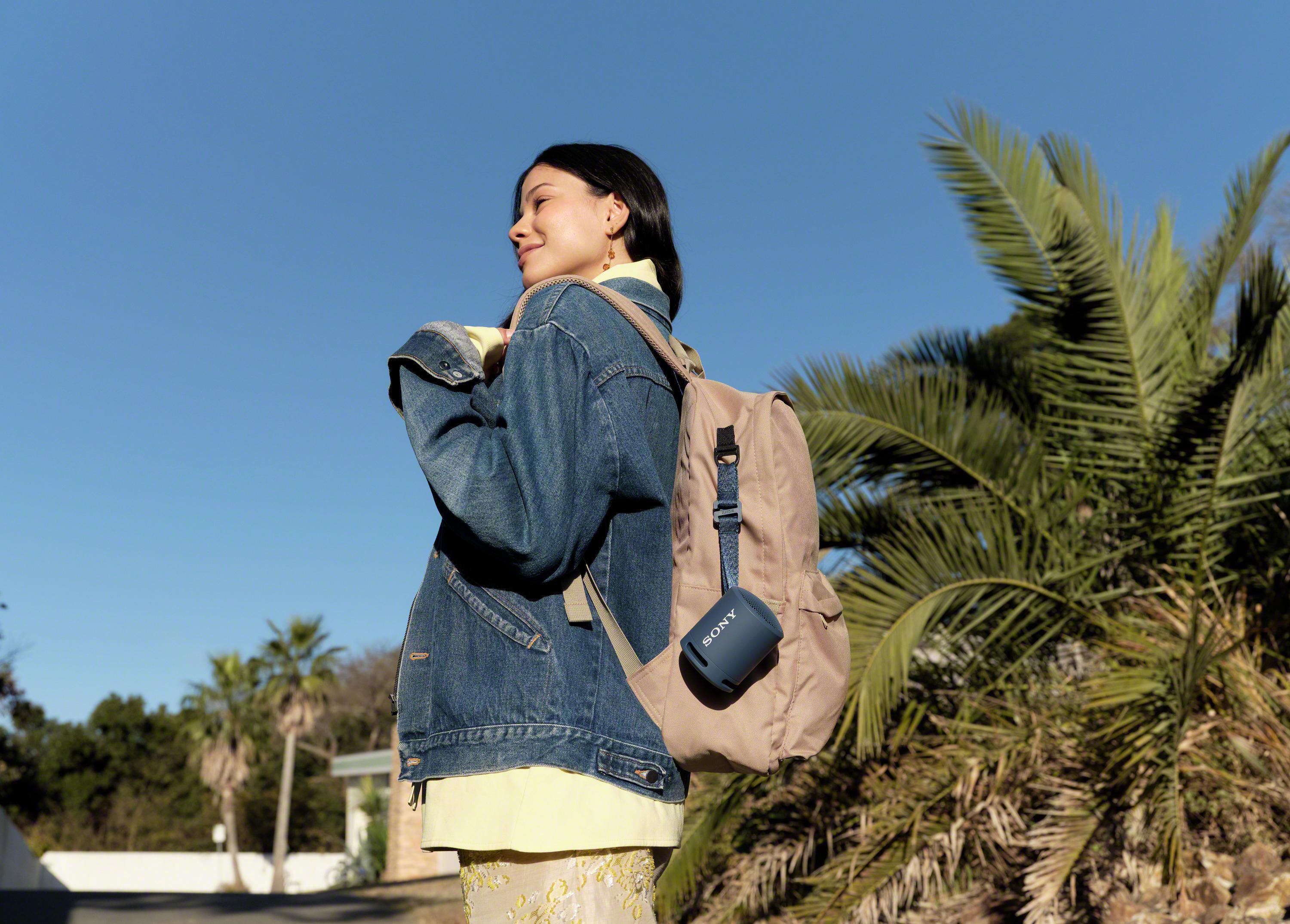 Frau mit Rucksack trägt einen marineblauen Sony-Lautsprecher, draußen mit blauem Himmel.