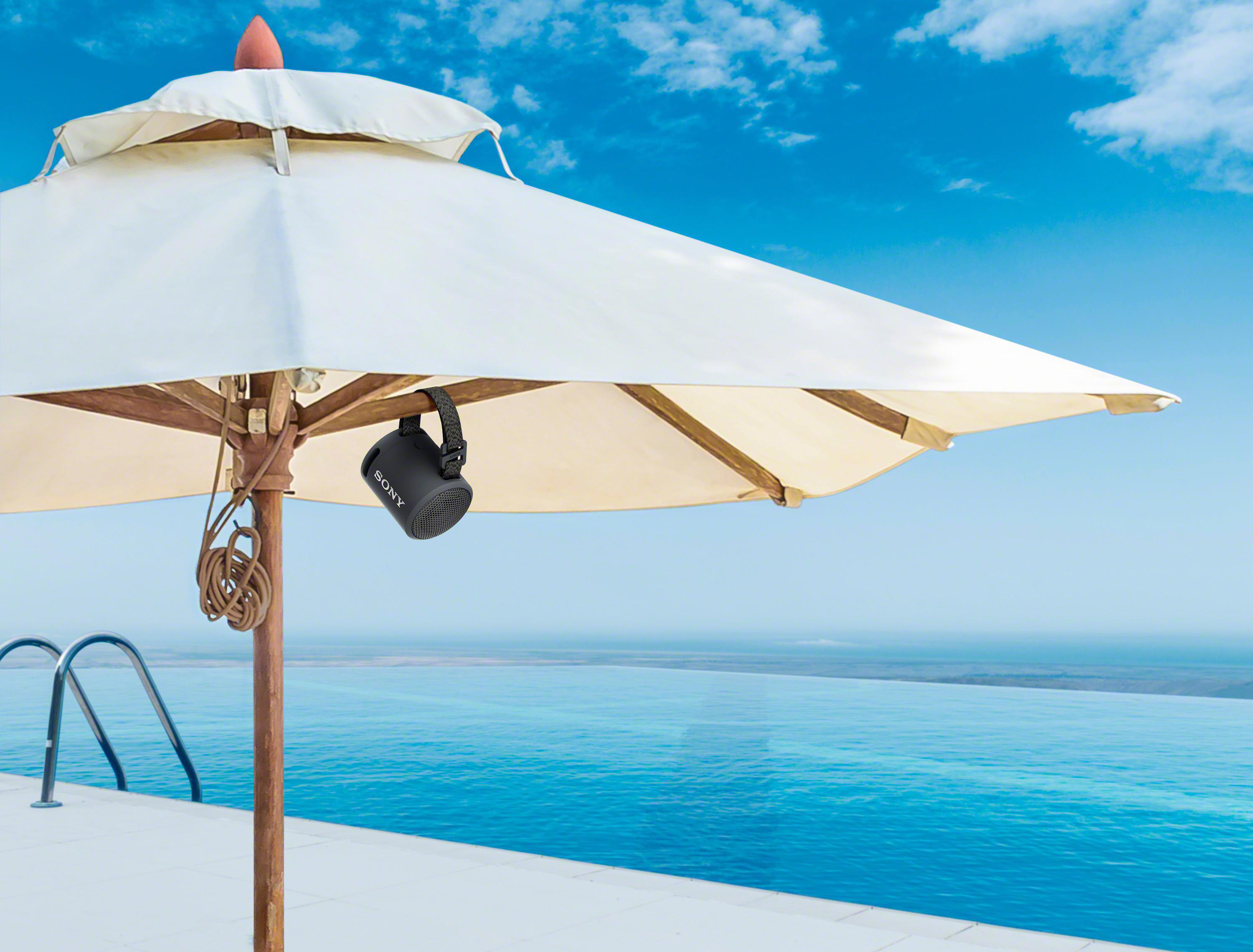 Schwarzer Lautsprecher hängt an weißem Sonnenschirm über Pool, blauer Himmel im Hintergrund.