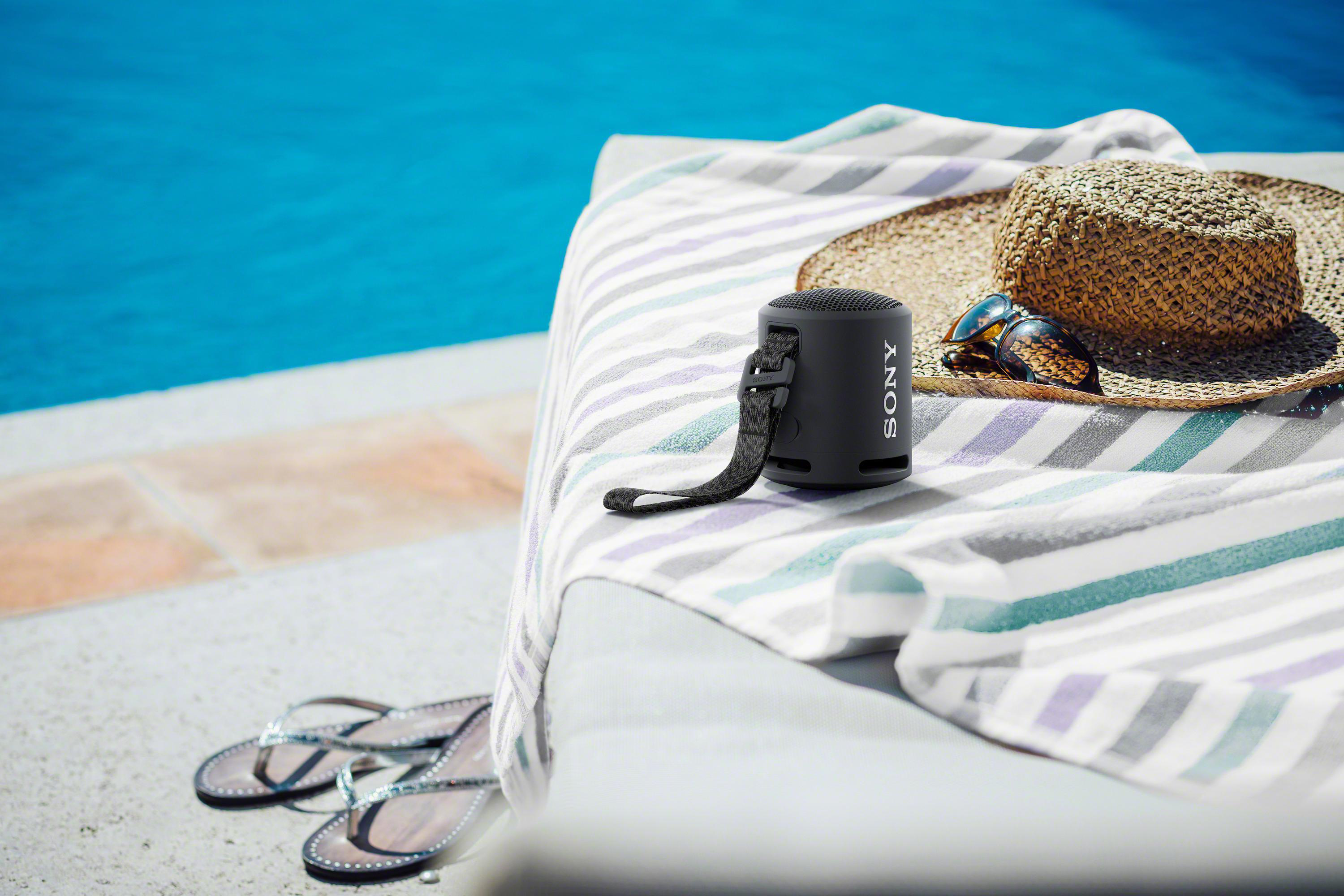 Schwarzer Sony-Lautsprecher auf Handtuch am Pool, Strohhut und Sonnenbrille daneben, Flip-Flops im Vordergrund.