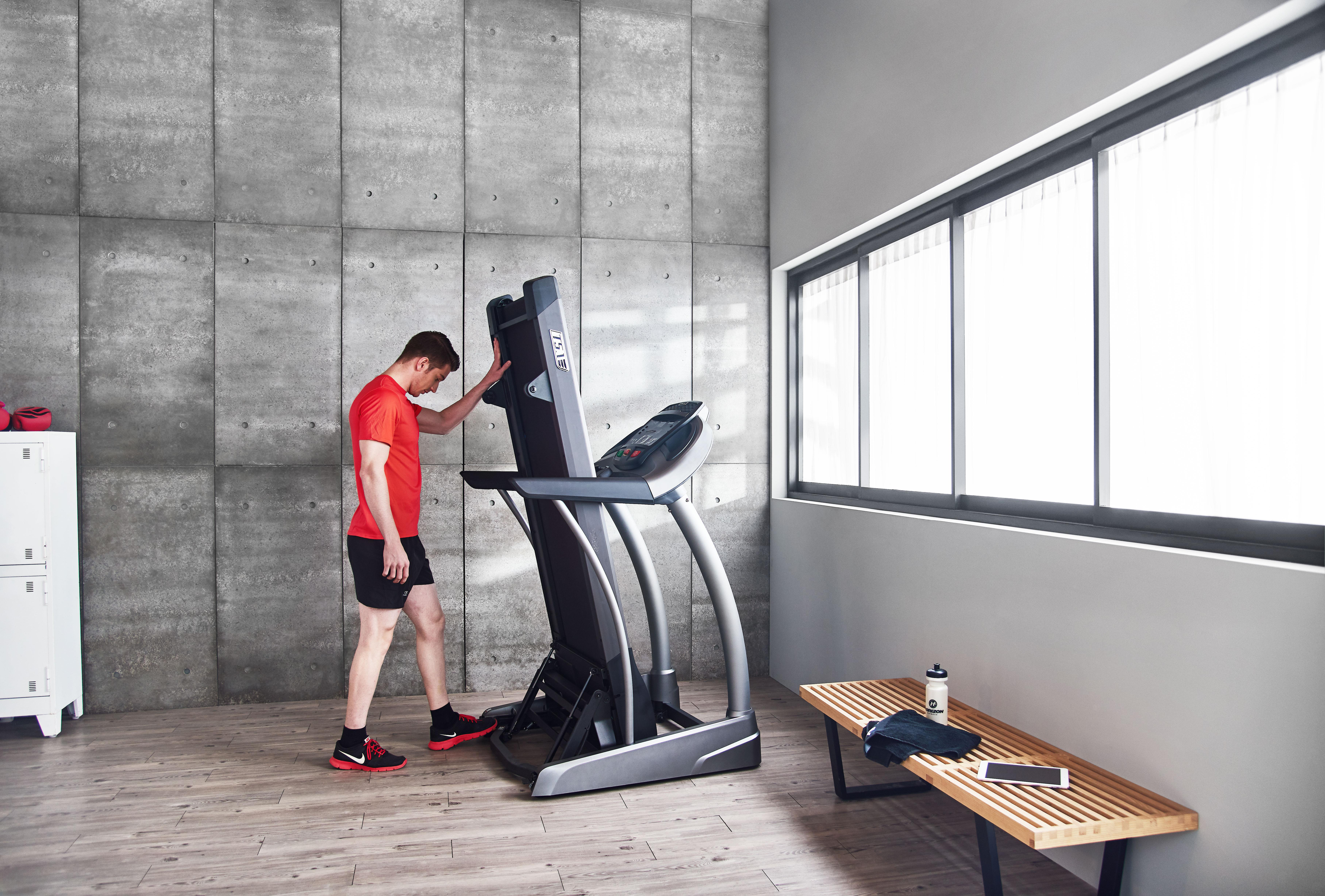 Ein Mann in rotem Hemd steht neben einem Laufband in einem Fitnessstudio und schaut nach unten.