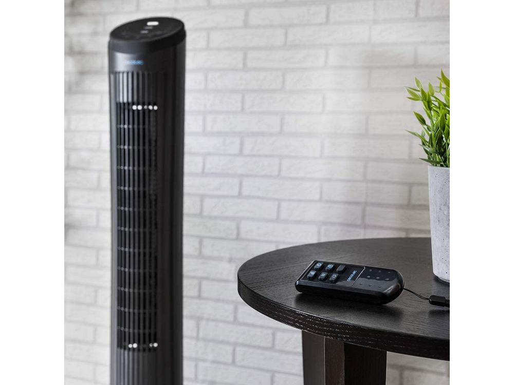 Ventilador de torre negro y mando a distancia en una mesa, con fondo de pared de ladrillo.