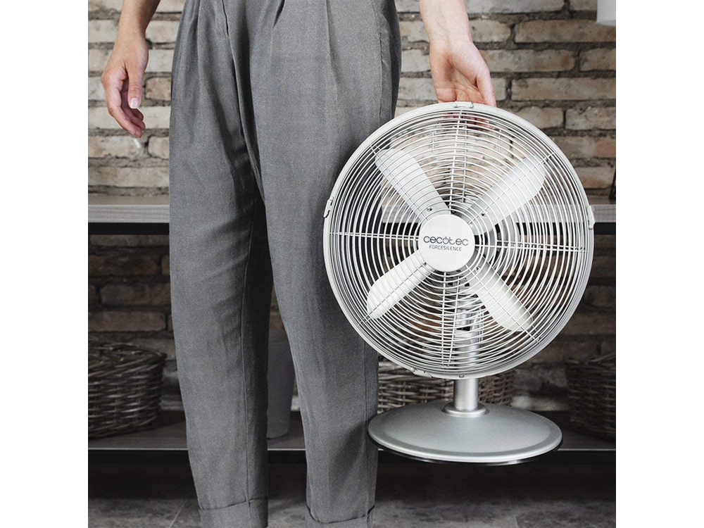 Una persona sosteniendo un ventilador blanco con cuatro aspas. La persona lleva pantalones grises, frente a una pared de ladrillo.