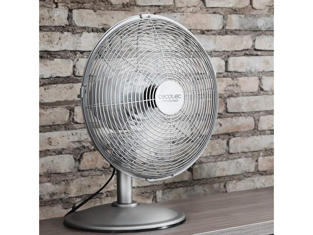 Ventilador de escritorio plateado, sobre una superficie de madera frente a una pared de ladrillos.