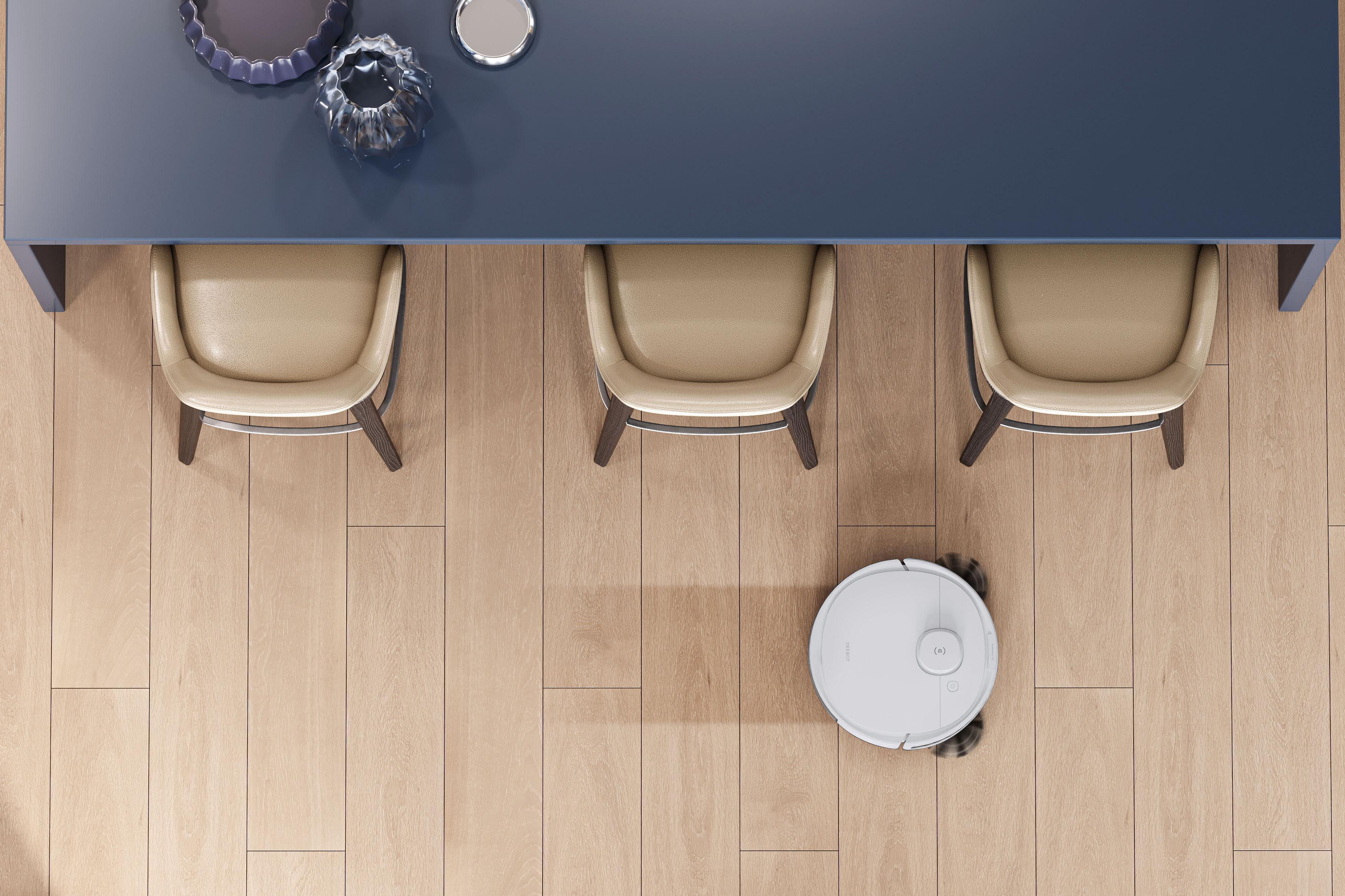 Draufsicht auf einen Essbereich: Tisch, drei Stühle und ein Roboter-Staubsauger auf Holzboden.