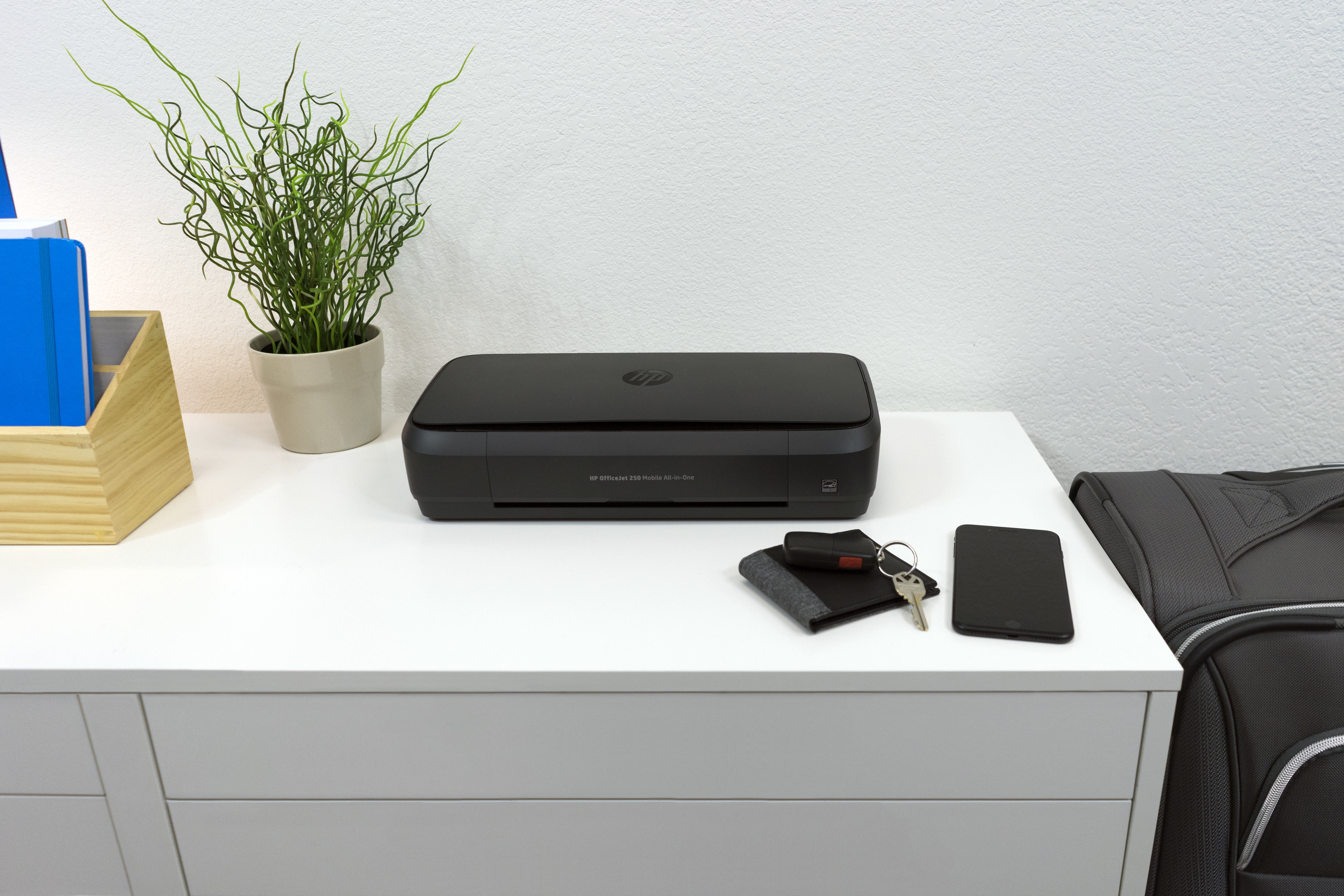 Una impresora HP, teléfono, cartera, llaves y una planta en un escritorio blanco.