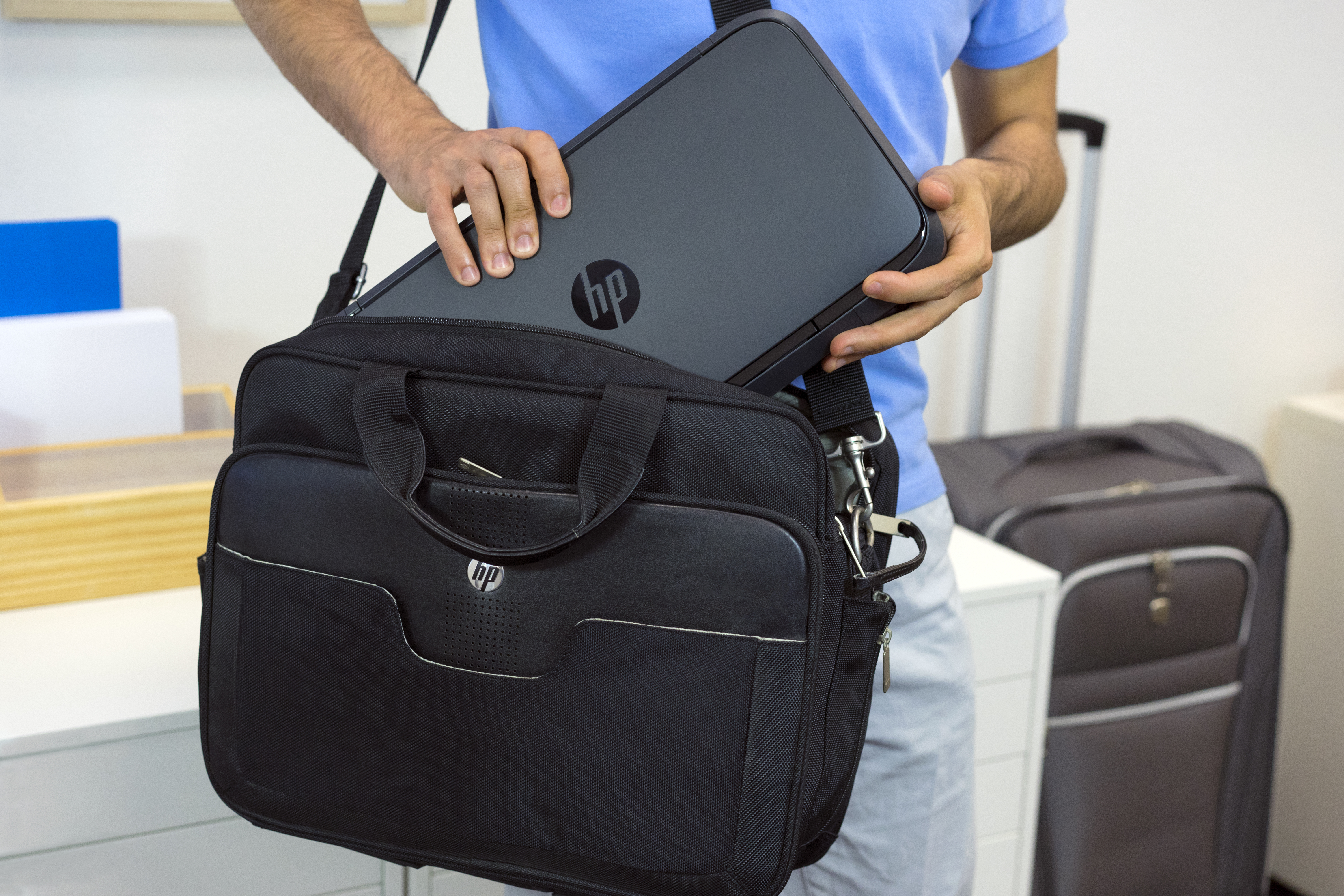 Persona metiendo una impresora HP negra en una bolsa negra, con una maleta en el fondo.
