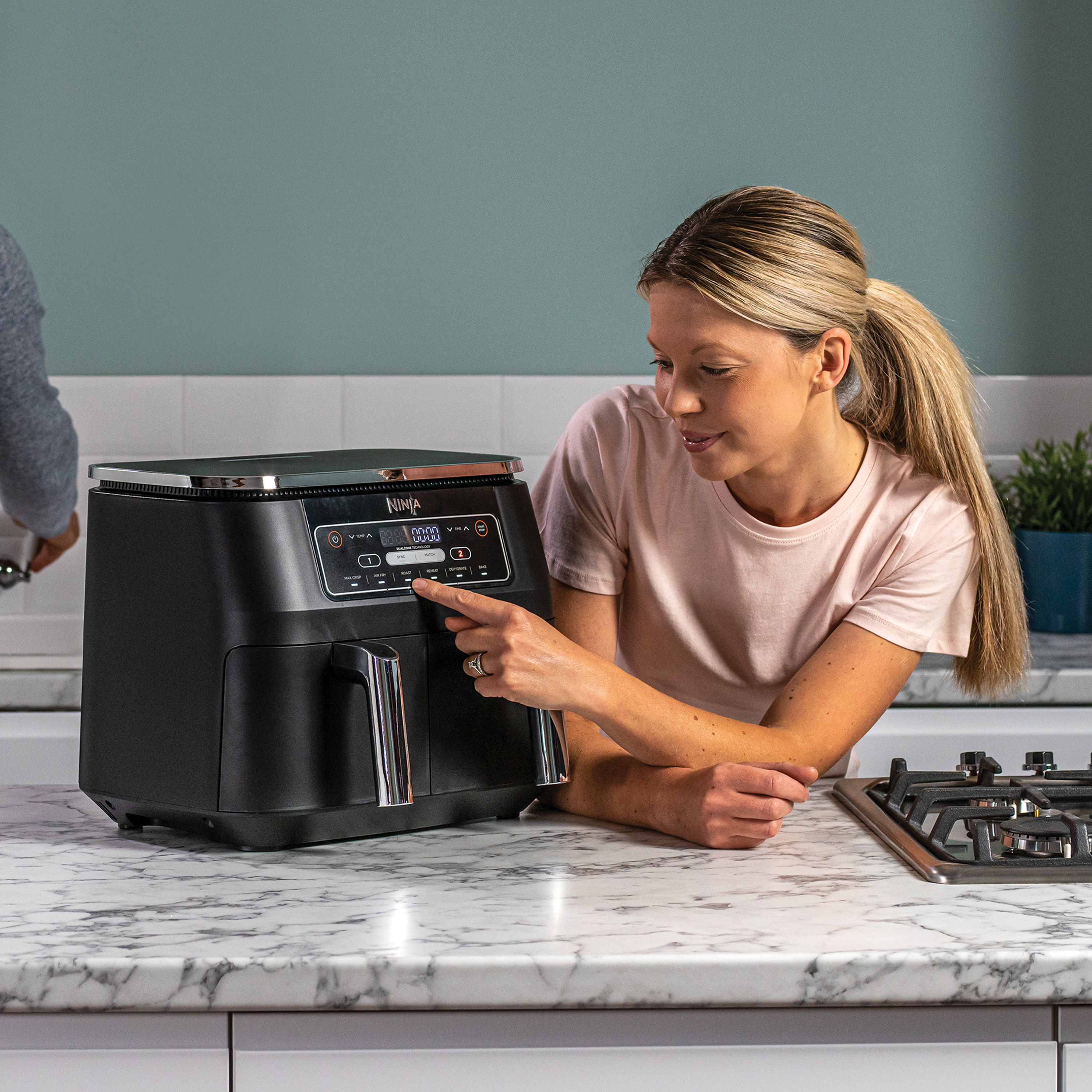Een vrouw die een zwarte Ninja airfryer gebruikt op een marmeren aanrecht in een keuken.