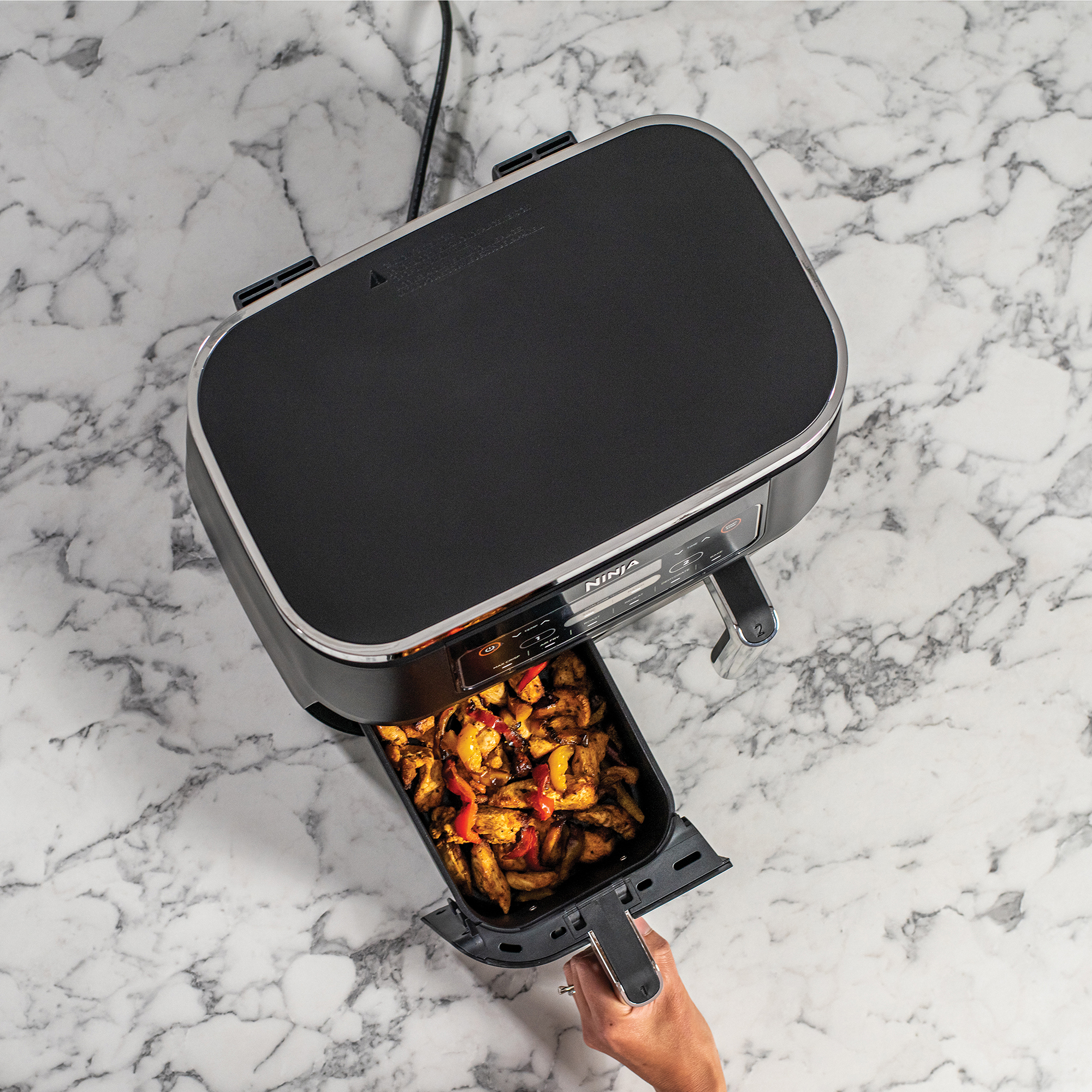 Een Ninja airfryer met een bak vol gekookt voedsel. De bak wordt eruit getrokken. Marmeren oppervlak.