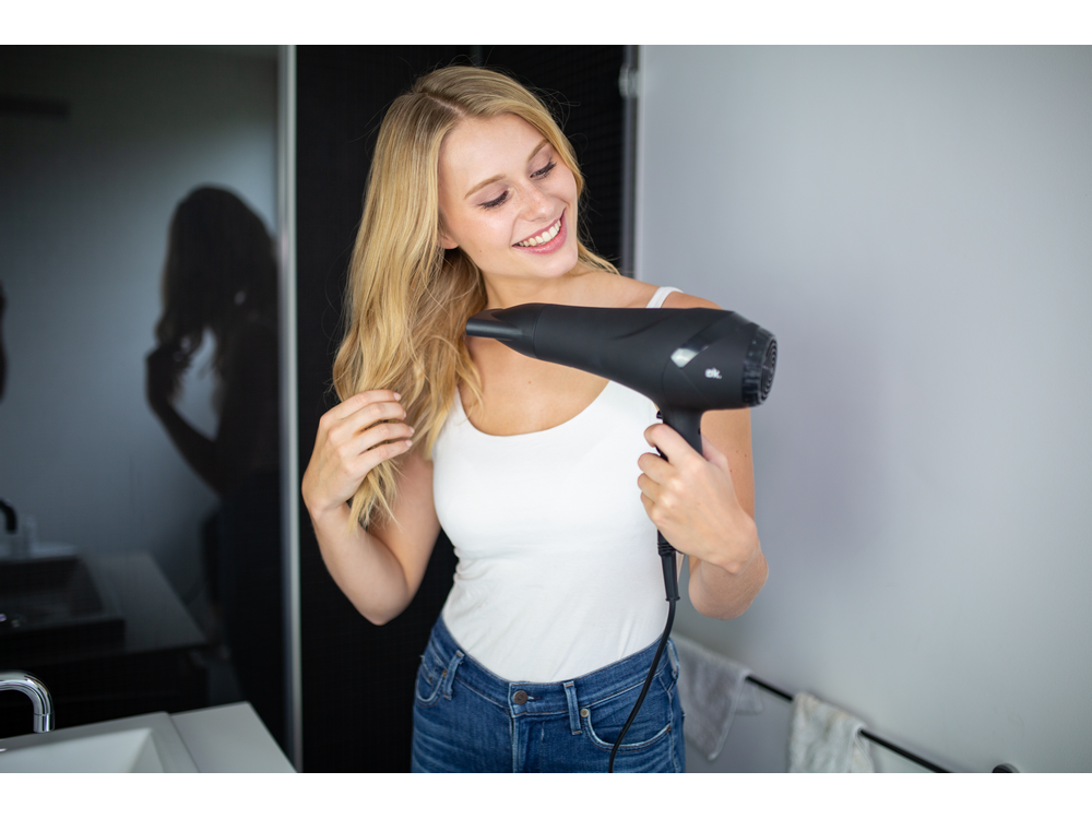 Mujer rubia secándose el pelo con un secador negro. Ella está sonriendo. Ambiente de baño.