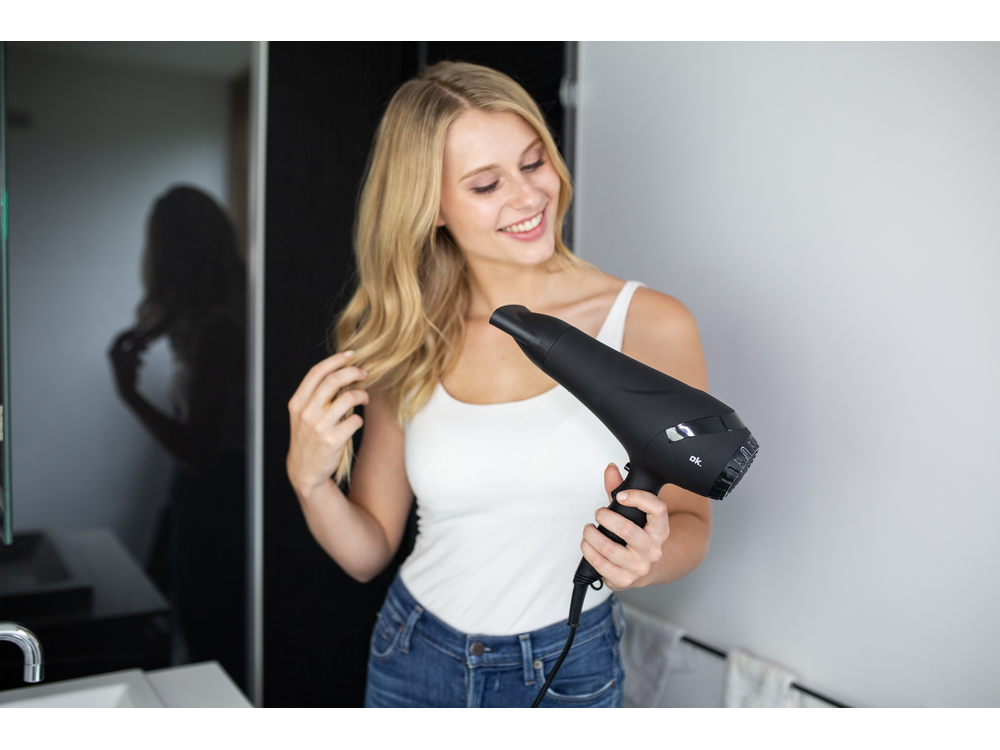 Mujer rubia sonriendo mientras sostiene un secador de pelo negro en un baño.