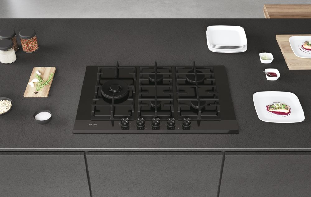 Una encimera de gas Haier negra sobre una encimera gris, con platos y guarniciones.