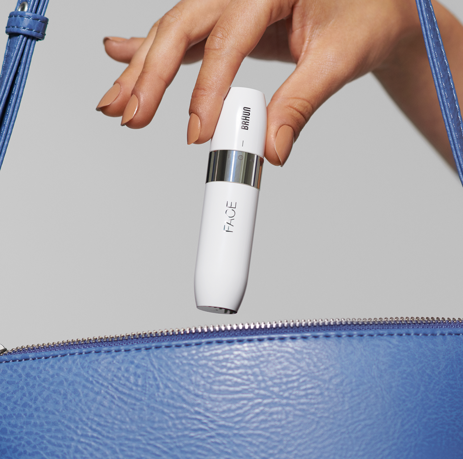 A hand holds a white Braun Face device near a blue purse. The background is a grey wall.