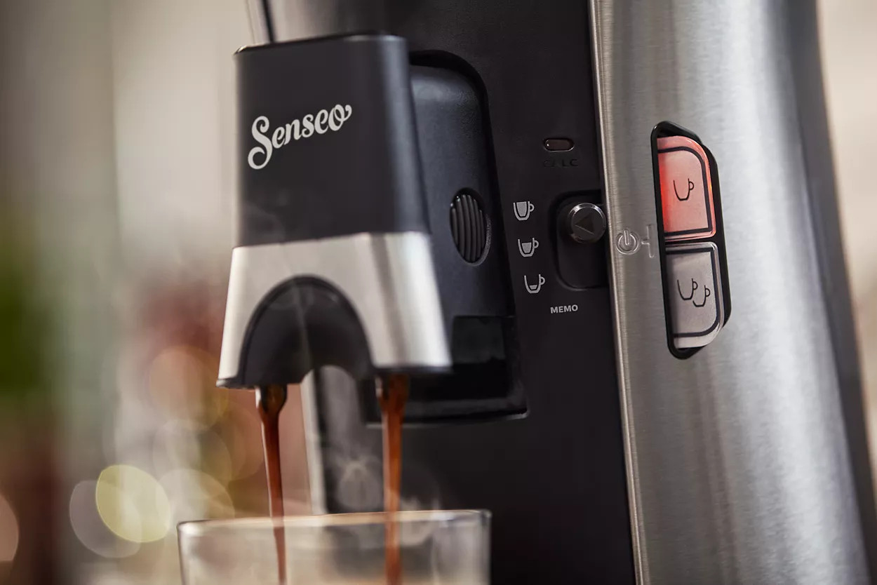 Un gros plan sur une machine à café avec du café qui se verse dans un verre et le logo 'Senseo'.