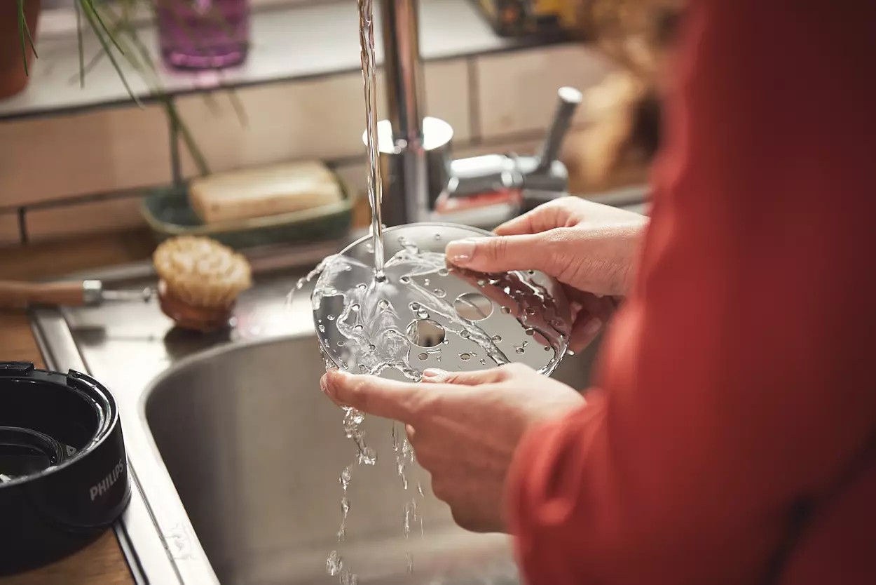 A person washing a food processor attachment under a running faucet in a kitchen sink, with a blurred background.