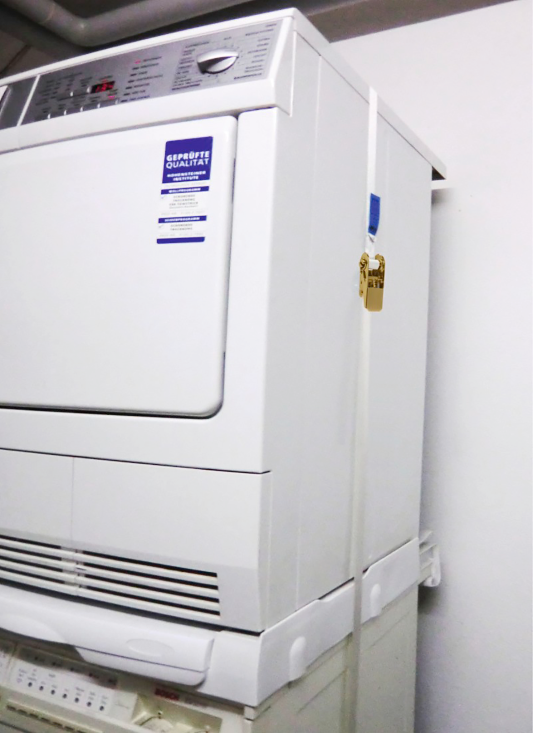 A white washing machine and dryer are stacked. A gold padlock secures the dryer to the washer.