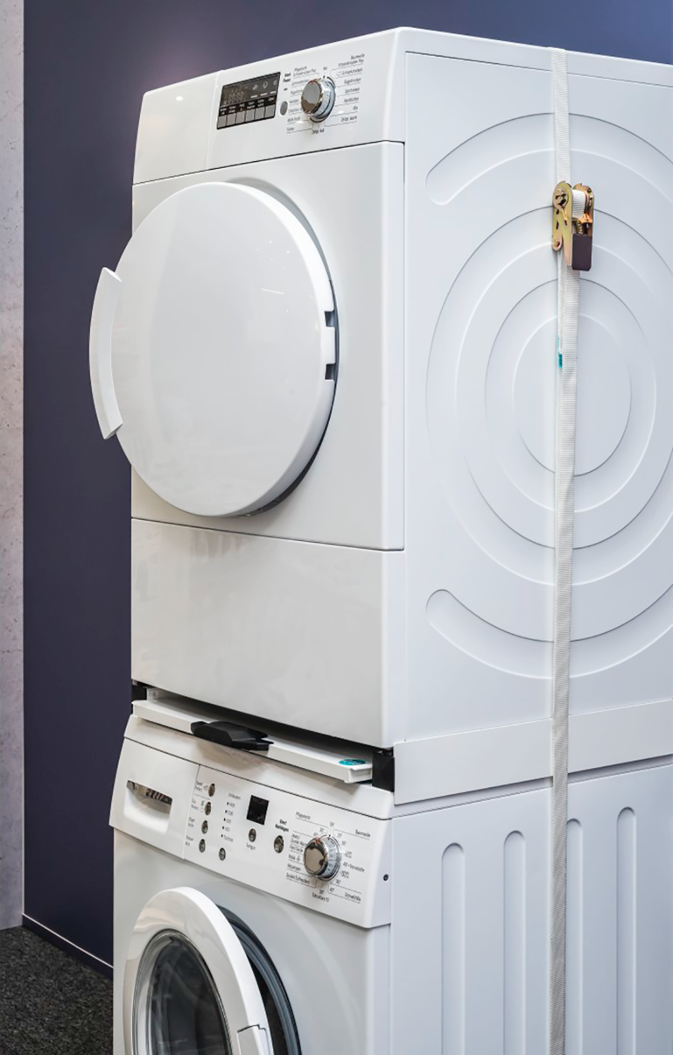 A stacked white washer and dryer set with open doors, secured with a strap against a blue wall.