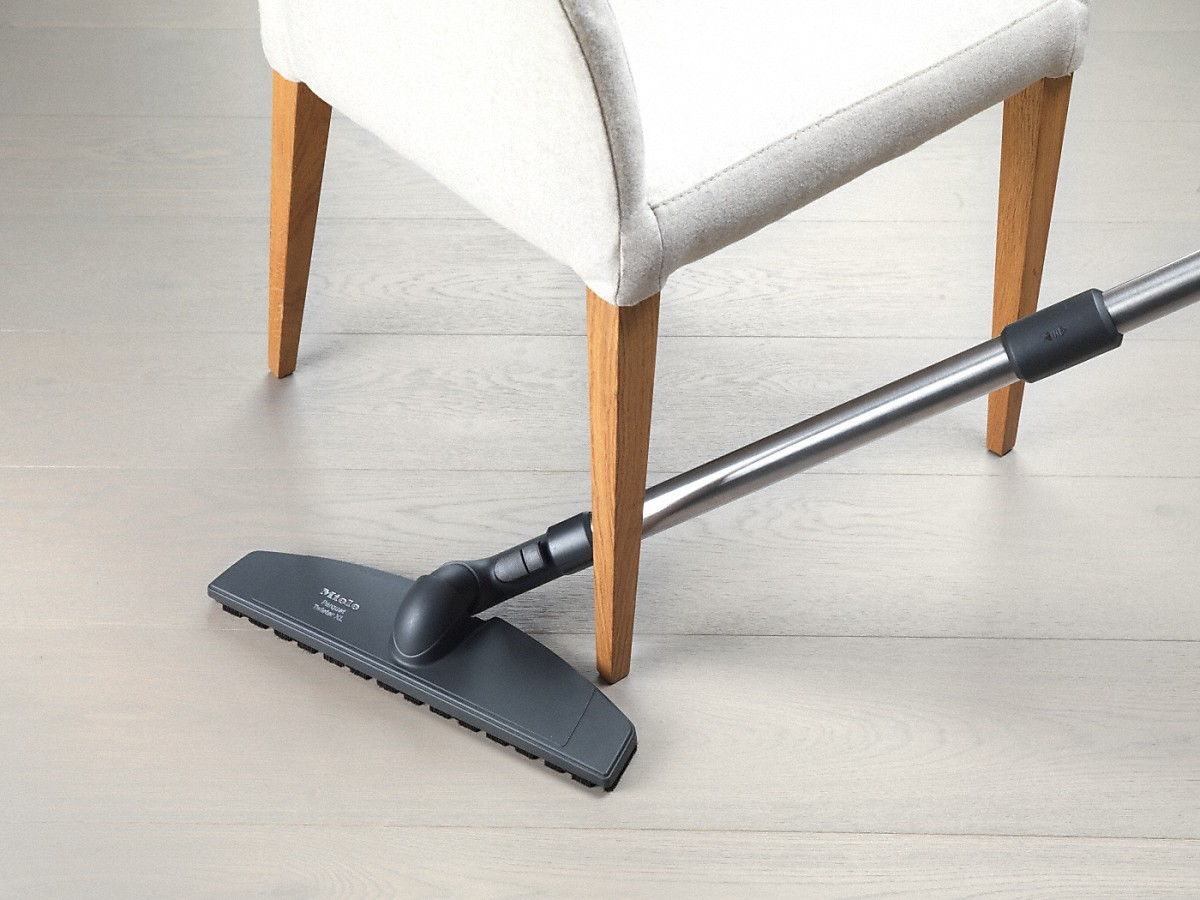 Stofzuigerkop die onder een stoel schoonmaakt. De stoel is wit met houten poten.
