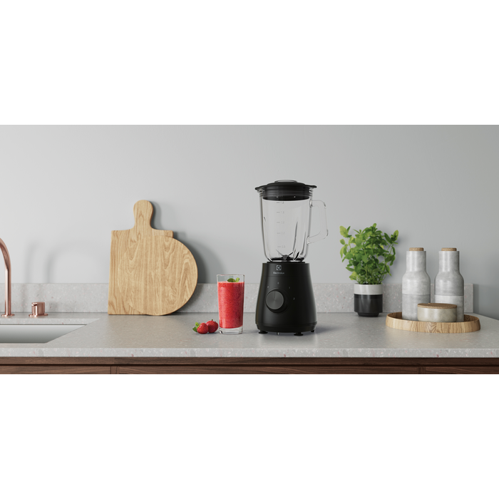 Fekete turmixgép egy pohár piros smoothie-val és egy fa vágódeszkával.