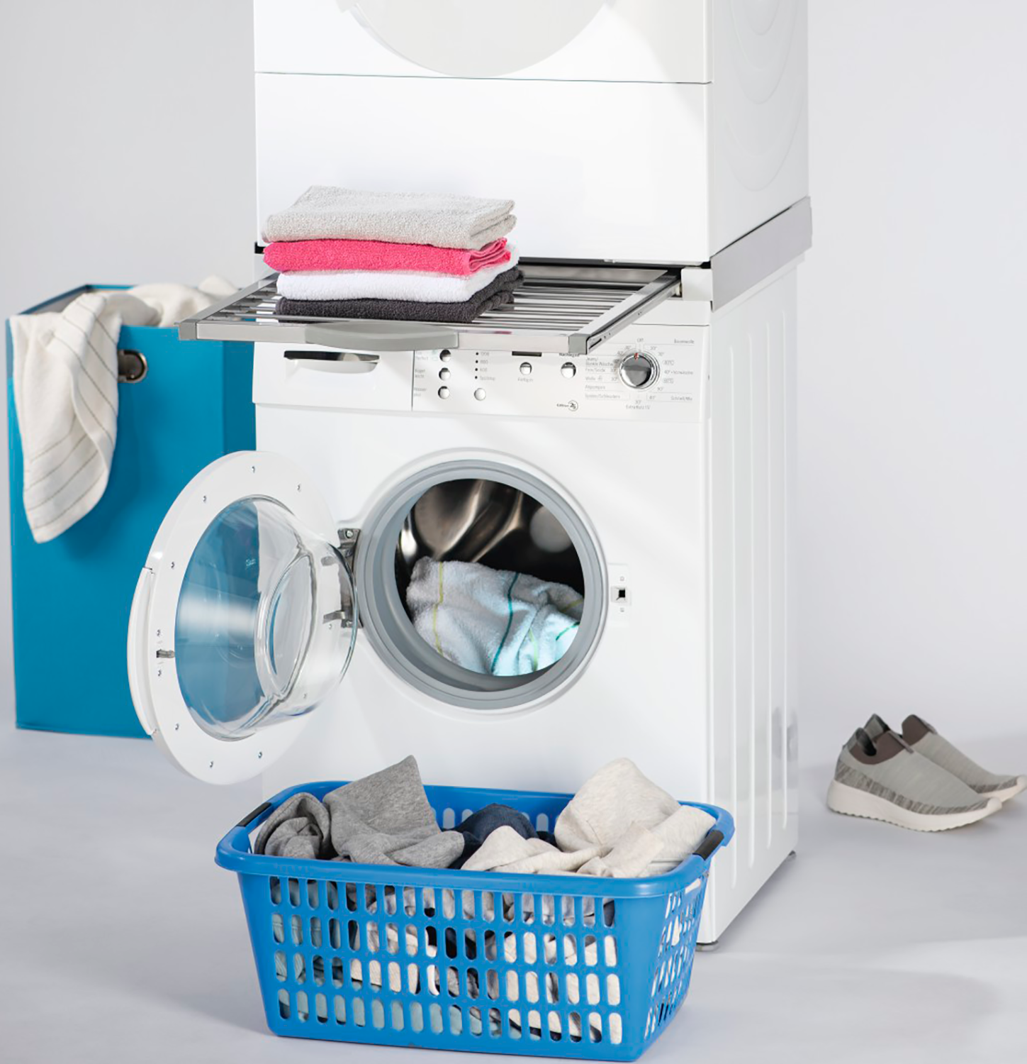 Een wasmachine met een open deur, een wasmand en een stapel handdoeken op een plank.