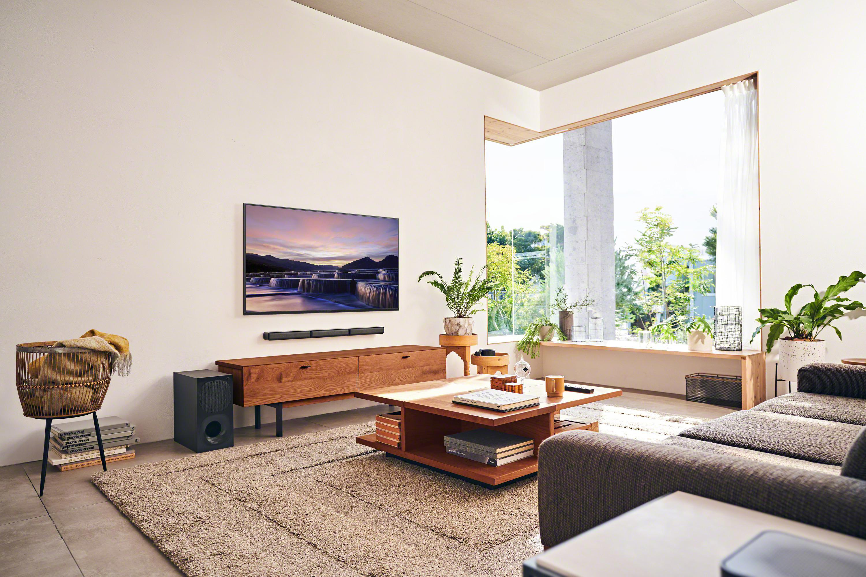 Ein modernes Wohnzimmer mit Fernseher, Soundbar und Subwoofer. Ein Fenster bietet einen Blick nach draußen.