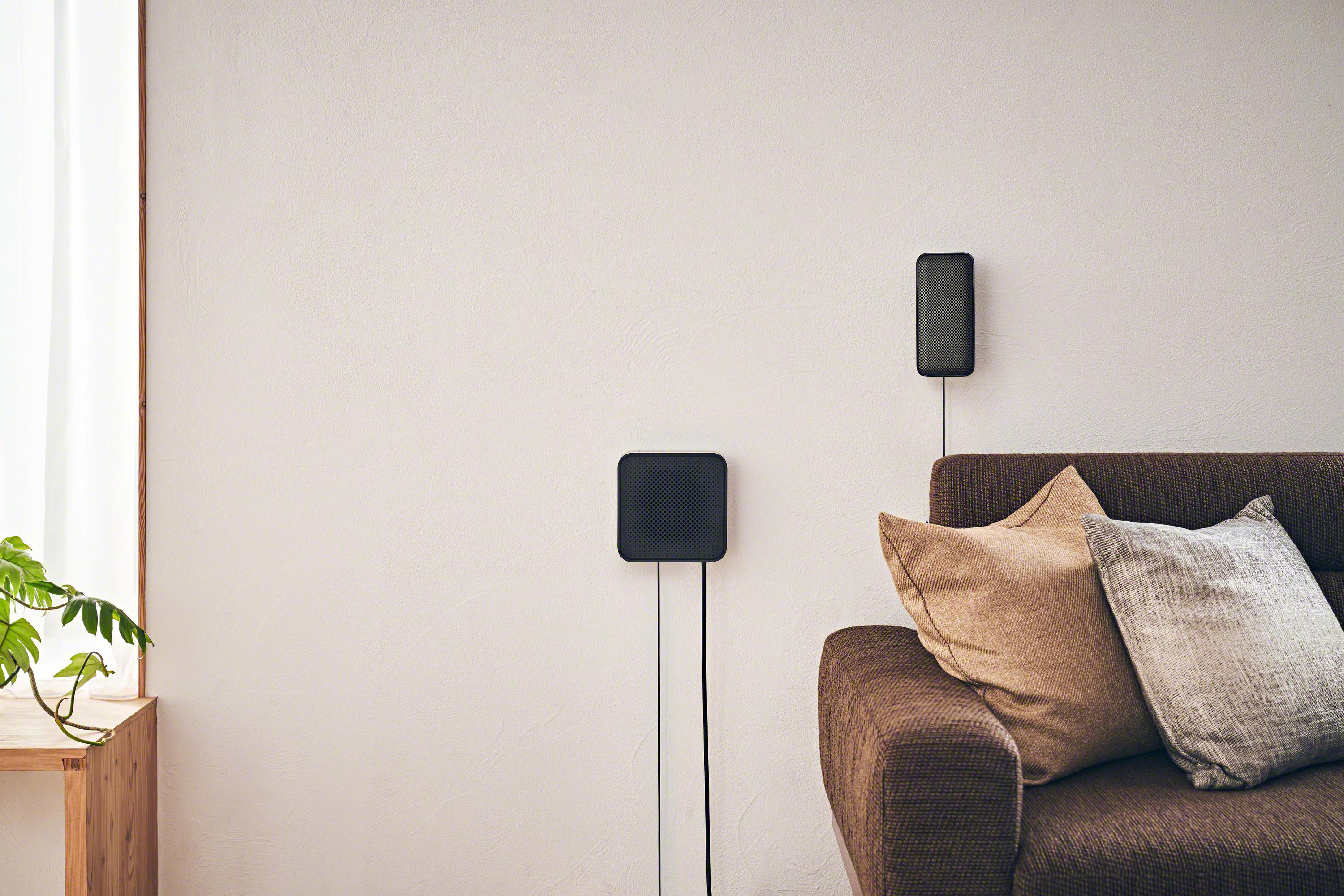 Ein modernes Wohnzimmer mit zwei Lautsprechern an der Wand und einem braunen Sofa. Links ist eine Pflanze.