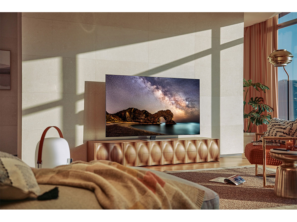 Un televisor muestra una escena de paisaje. Una estantería de madera, un sofá, una alfombra y una lámpara están en primer plano.