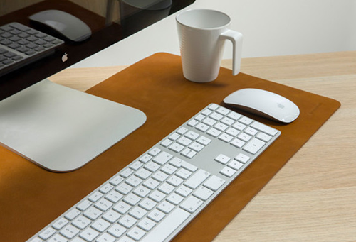 A desk setup with a white keyboard, mouse, and mug on a brown leather mat. A computer monitor is in the background.