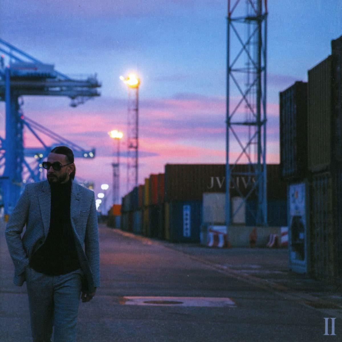Ein Mann in Sonnenbrille und grauem Anzug geht auf einem Dock. Gebäude und Container befinden sich im Hintergrund.