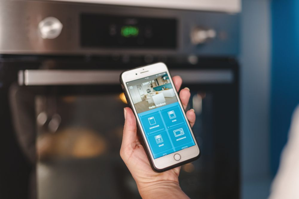 Una mano sosteniendo un teléfono que muestra una aplicación de hogar inteligente con iconos de horno y otros electrodomésticos.