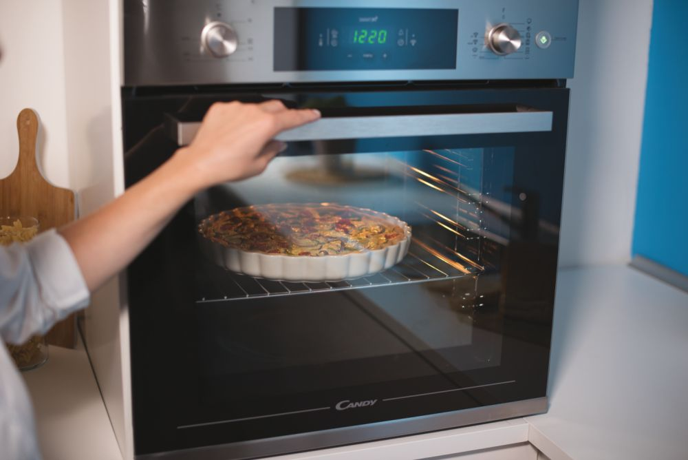 Una mujer abriendo la puerta del horno, revelando un pastel dentro. Una cocina blanca.