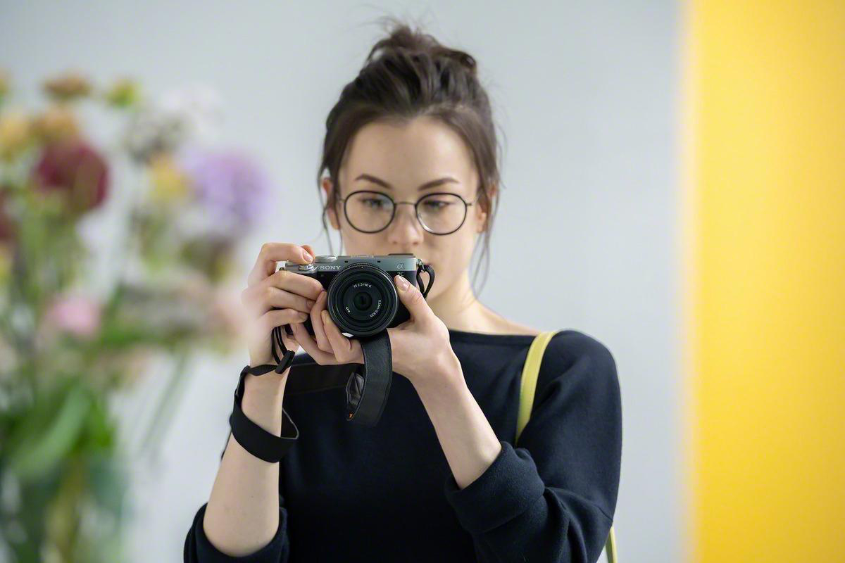Eine Frau mit Brille macht ein Foto mit einer Kamera. Eine gelbe Wand im Hintergrund.