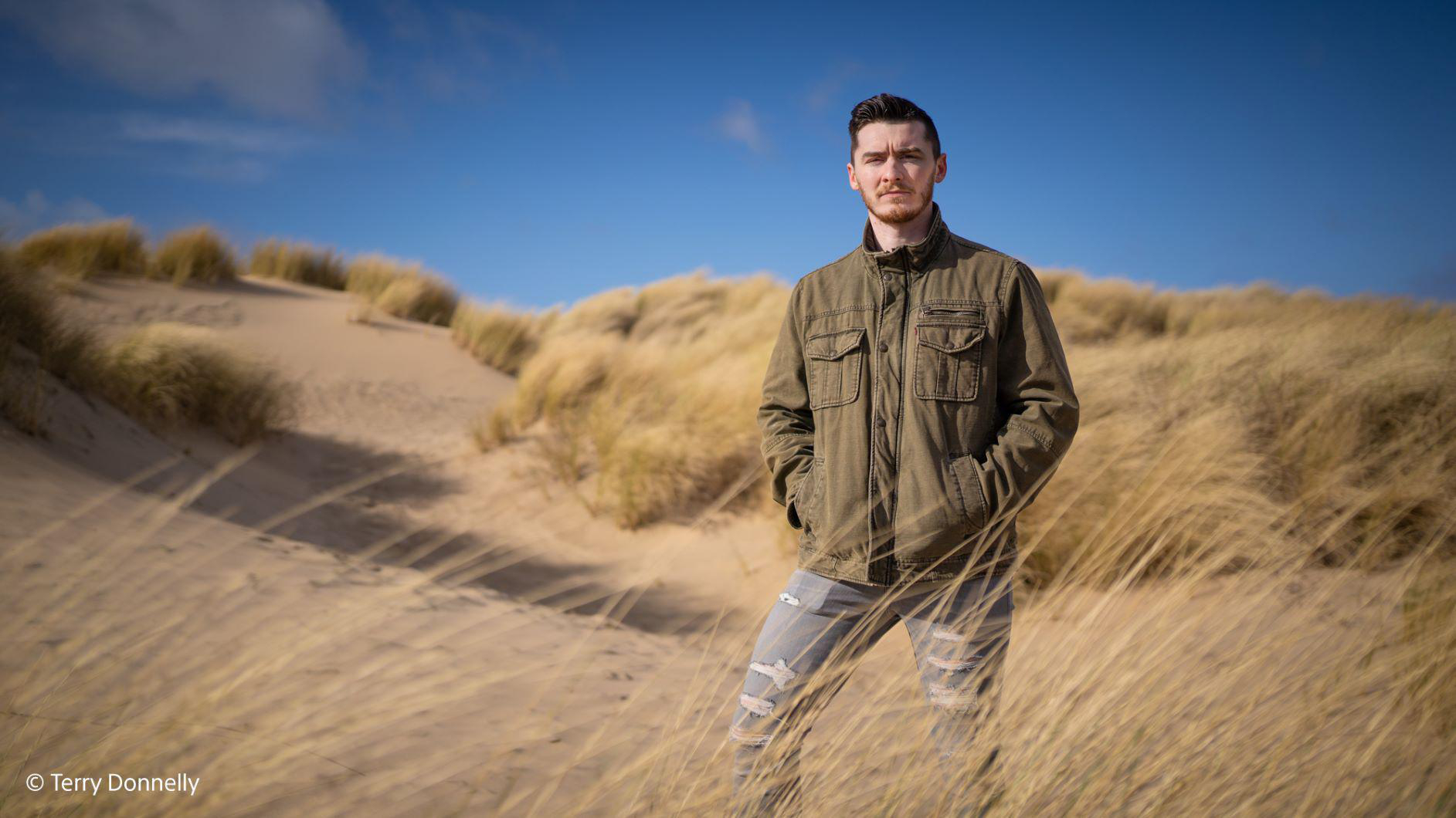 Ein Mann mit Bart steht in den Dünen. Er trägt eine Jacke und Jeans. Sonniger Tag.