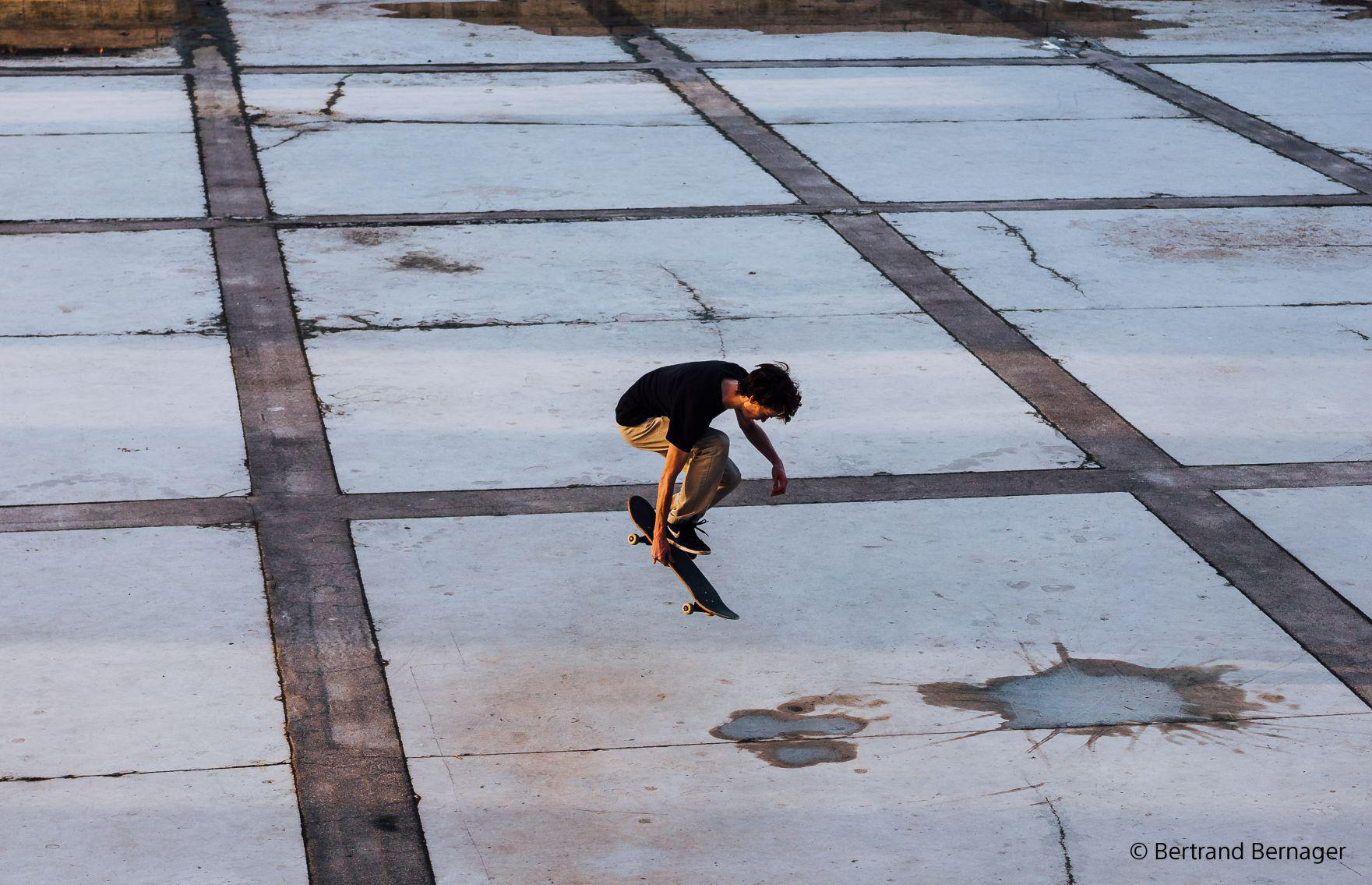 Ein junger Mann in der Luft auf einem Skateboard. Der Boden ist Beton mit Rissen und Flecken.