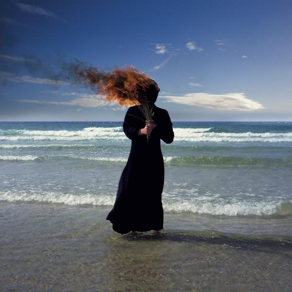 Eine Person in einem schwarzen Kleid steht im Wasser und hält ein Bündel Schilf. Der Himmel ist blau.