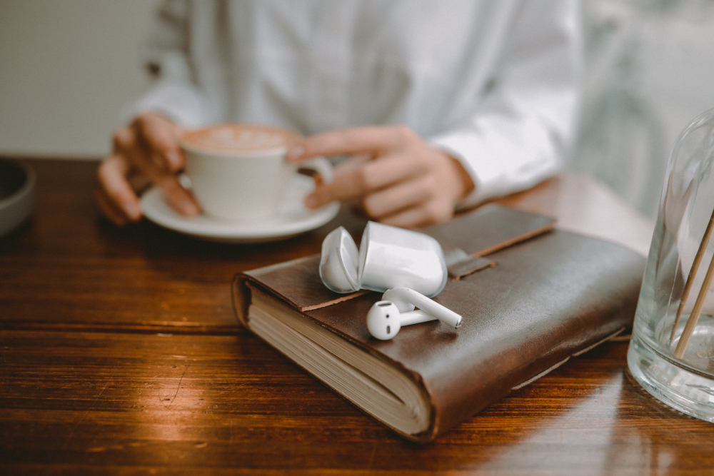 A person holding a coffee cup, with earbuds and a notebook on a wooden table.