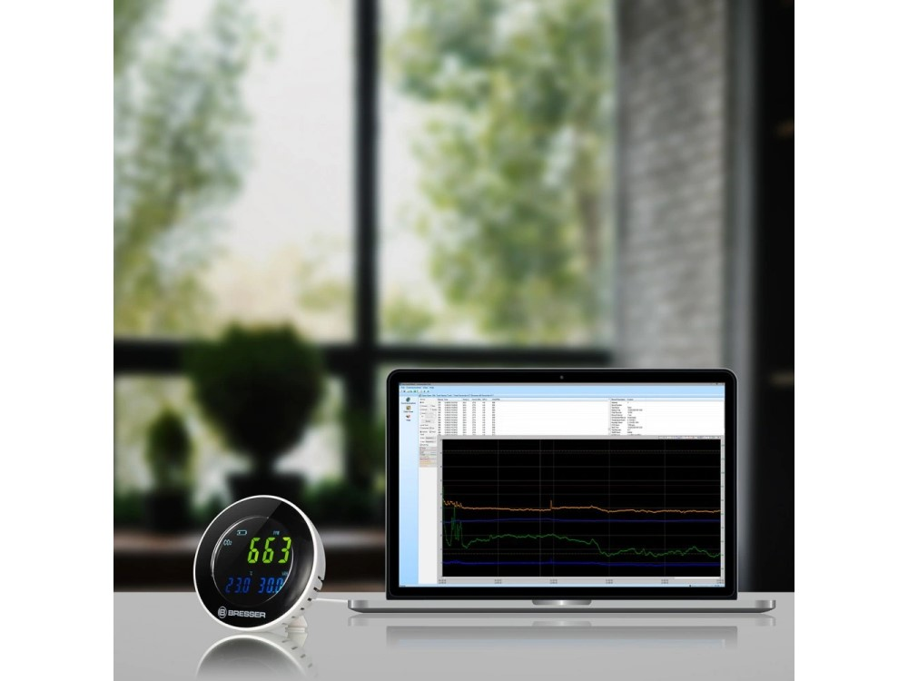 Medidor de CO2 junto a una laptop que muestra un gráfico. Ventana y vista exterior al fondo.