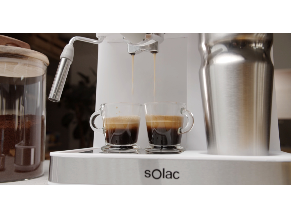 Máquina de espresso dispensando café en dos tazas de vidrio. Una jarra de granos de café y una jarra de metal están al lado.