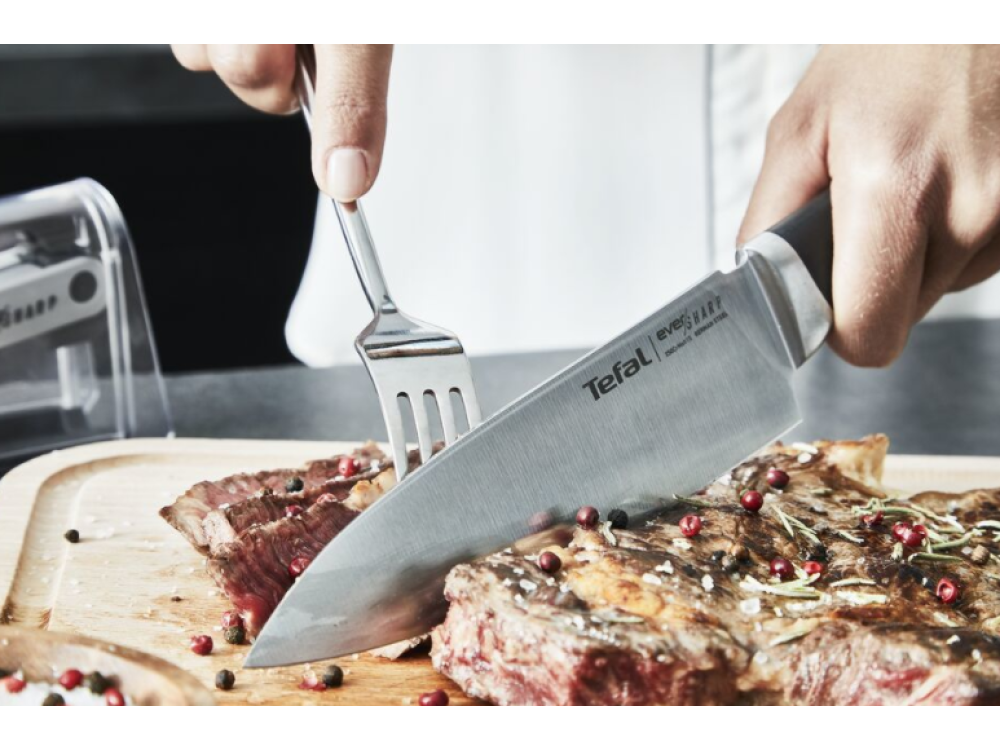 Una persona cortando un filete con un cuchillo y un tenedor sobre una tabla de cortar de madera con granos de pimienta.