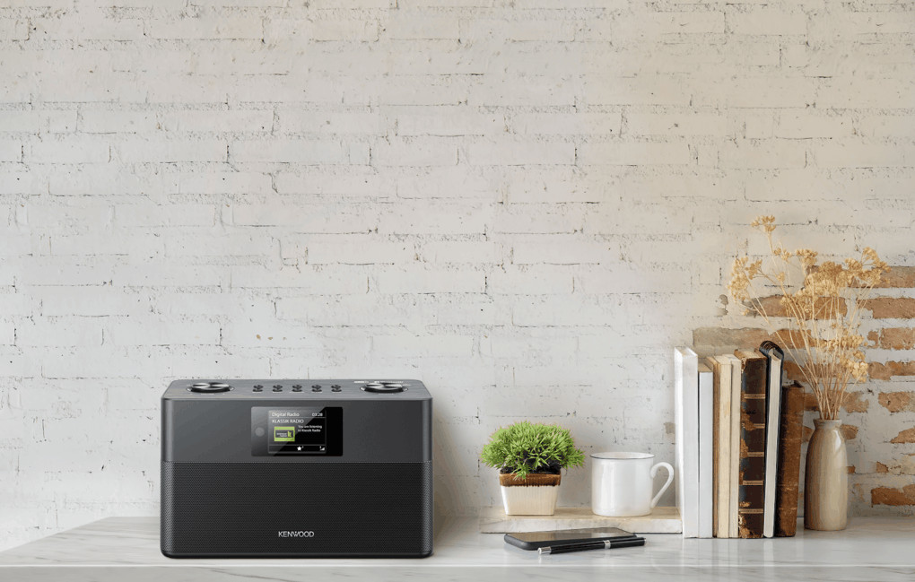 Een zwarte Kenwood-radio op een marmeren oppervlak, boeken, een plant en een mok voor een bakstenen muur.
