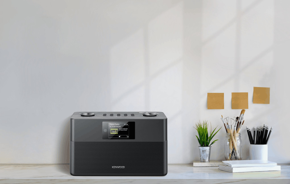Een zwarte Kenwood-radio op een wit bureau met planten en pennen tegen een witte muur met plaknotities.