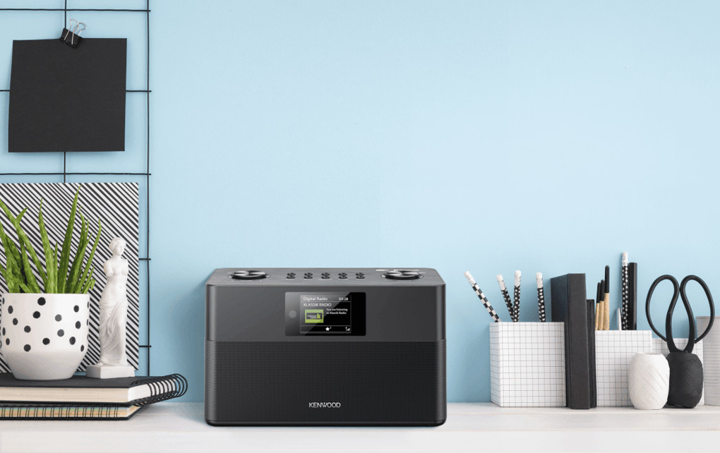 Een zwarte Kenwood-radio op een wit bureau. Er staat een plant, pennen en een notitieblok op het bureau.