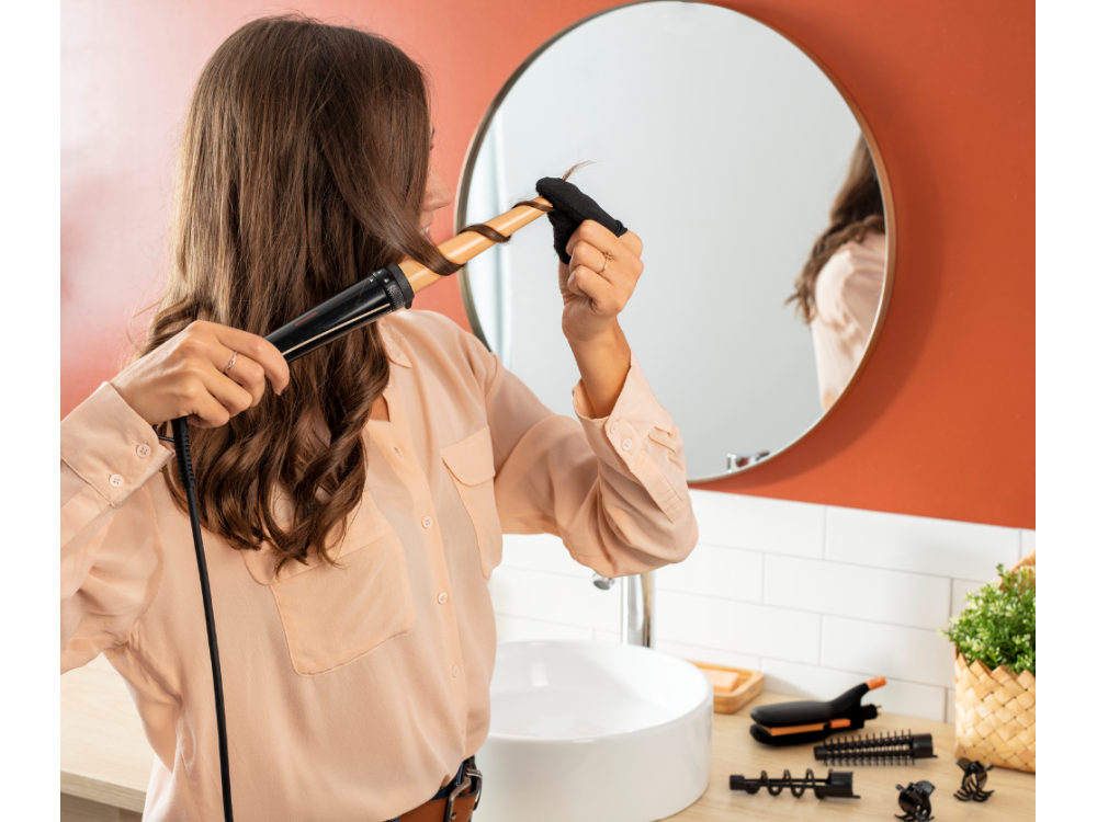 Mujer se riza el pelo con un rizador en un baño, espejo redondo y lavabo al fondo.