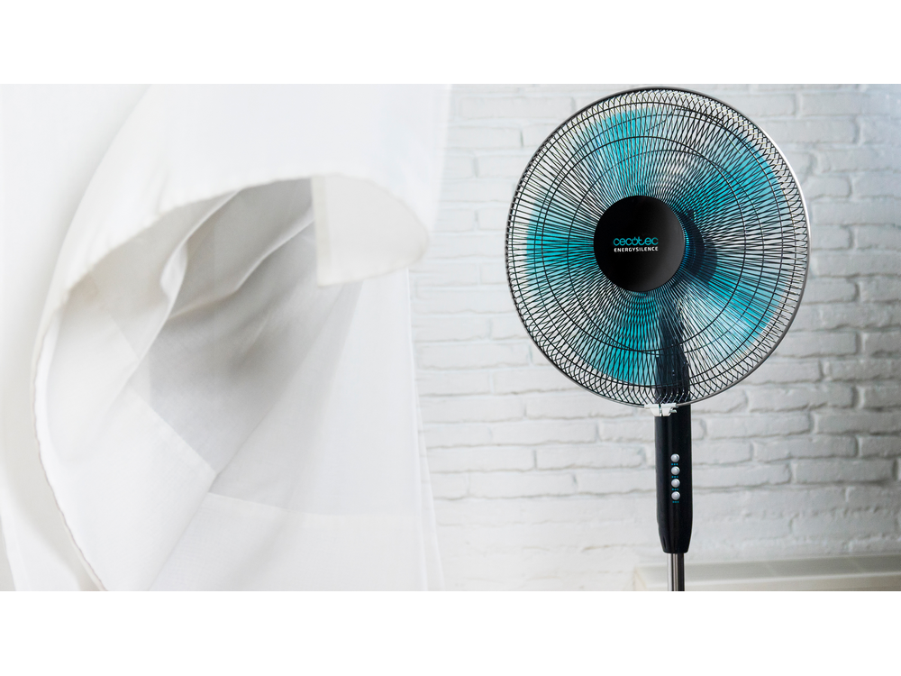 Ventilador de pie negro y azul con tela blanca en el fondo. Se puede ver una pared de ladrillo.