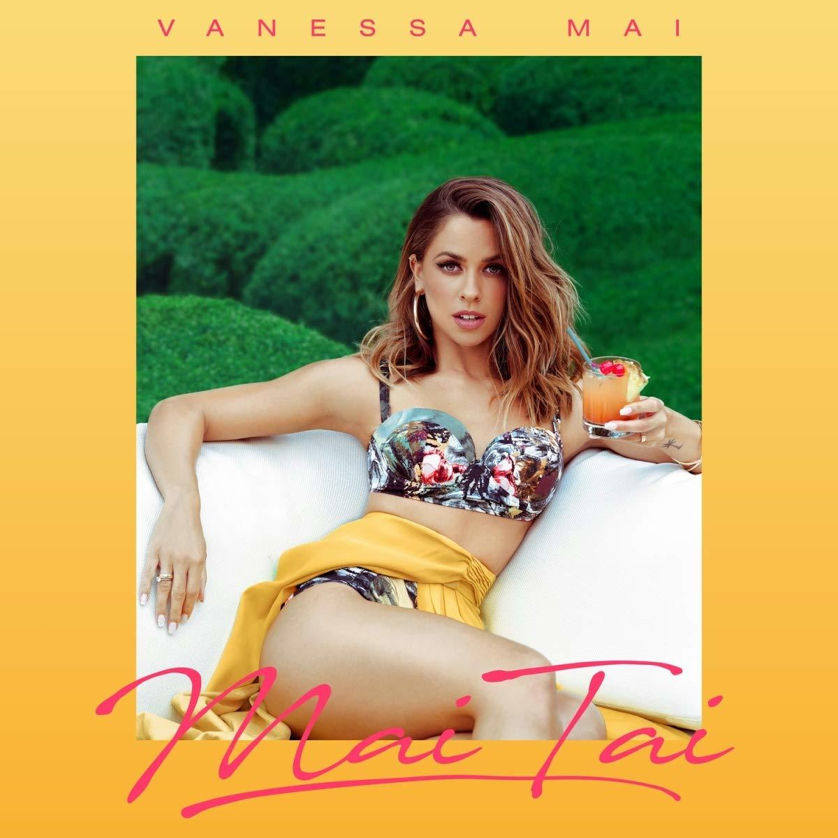 Vanessa Mai auf einer weißen Couch, mit Cocktail, Blumen-Top und gelbem Rock. Grüner Hecken-Hintergrund, pinkfarbenes 'Mai Tai'.