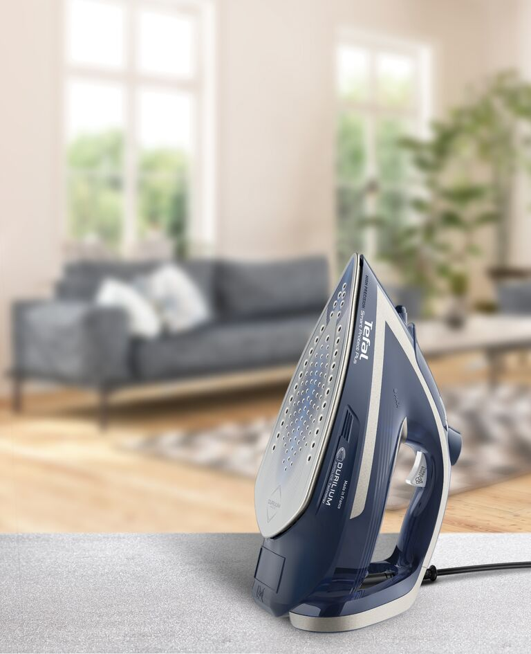 Een blauw en wit strijkijzer op een tafel. Een wazige woonkamer op de achtergrond.
