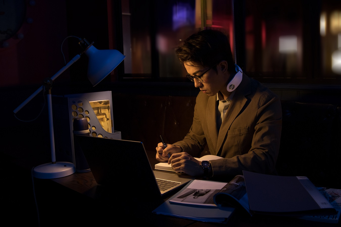 Person arbeitet an einem Schreibtisch, trägt ein Nackenband, Laptop und Lampe. Dunkle Raumeinstellung, schreibt.