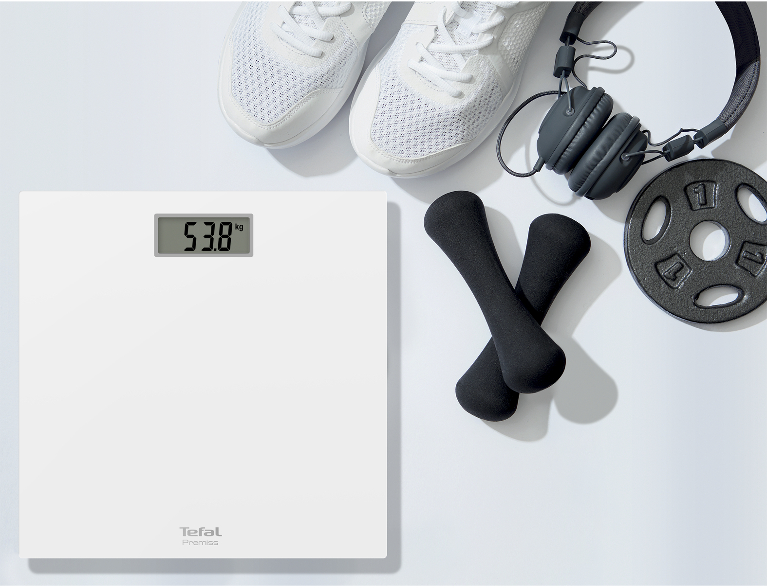 Witte weegschaal toont 53,8kg. Sneakers, koptelefoon, dumbbells en gewichtschijf op witte achtergrond.