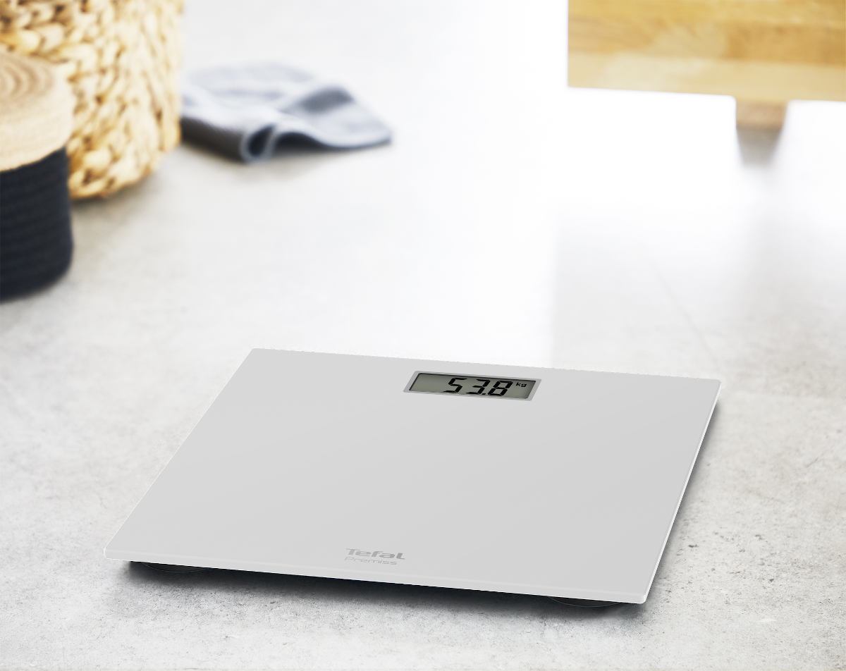 Een witte digitale weegschaal geeft 53,8 kg weer. Een gevlochten mand, grijze handdoek en hout in de vage achtergrond.
